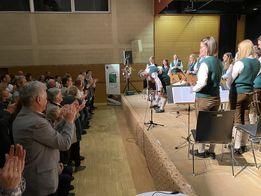 Eine Gruppe Musiker tritt auf der Bühne in einem Saal auf, während eine Menge zuklatscht und zusieht. Die Musiker spielen verschiedene Instrumente, und der Saal ist gut beleuchtet.