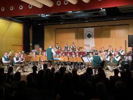 Ein Blasorchester spielt auf der Bühne vor einem Publikum. Die Musiker sind in traditioneller Kleidung gekleidet. Die Bühne ist mit verschiedenen Musikinstrumenten ausgestattet, und das Publikum sitzt davor.