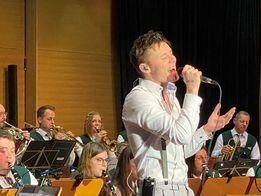 Ein Mann in einem weißen Anzug singt in ein Mikrofon und tritt vor einer Band auf. Die Bandmitglieder spielen verschiedene Instrumente, und das Publikum schaut zu. Die Umgebung scheint ein Konzert zu sein.