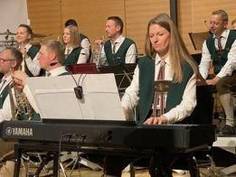 Eine Gruppe von Musikern tritt auf einer Bühne auf. Eine Frau spielt Klavier, während andere verschiedene Instrumente spielen, darunter Trompeten und Saxophone. Der Hintergrund ist eine verschwommene Wand mit Musikinstrumenten.