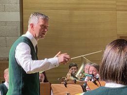 Ein Mann dirigiert eine musikalische Aufführung, hält einen Taktstock in der rechten Hand, während andere Instrumente spielen und im Hintergrund singen.