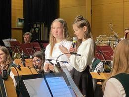 Zwei junge Mädchen stehen auf der Bühne, halten Mikrofone und singen vor einem Orchester. Andere Musiker spielen hinter ihnen, und mehrere Menschen schauen zu.