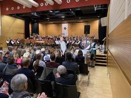 Ein Publikum sitzt in einem Saal und klatscht. Auf der Bühne spielt eine Gruppe von Musikern Instrumente. Ein Mann steht in der Mitte und spricht das Publikum an.