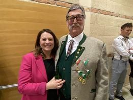 Ein Mann und eine Frau stehen nah beieinander und lächeln. Der Mann trägt ein traditionelles deutsches Outfit mit Medaillen. Dahinter steht ein Mann in einem weißen Hemd und grauer Hose mit verschränkten Armen.