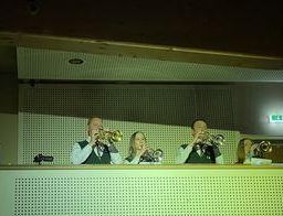 Vier Personen spielen Trompeten auf der Bühne; sie tragen Uniformen und sind von grünen Bühnenlichtern beleuchtet. Hinter ihnen befindet sich eine weiße Lochwand, eine Deckenleuchte und ein Notausgangsschild.