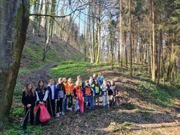 Eine Gruppe von Menschen versammelt sich in einem bewaldeten Gebiet, einige halten Taschen, und sie lächeln. Der Boden ist mit Blättern und kleinen Pflanzen bedeckt, und die Bäume sind hoch und blattreich.