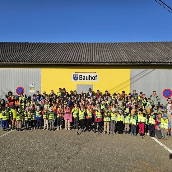 Eine große Gruppe von Kindern und Erwachsenen in Warnwesten posiert für ein Foto vor einem Gebäude mit dem Wort Bauhof darauf.