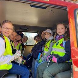 Eine Gruppe von Kindern in Warnwesten und Handschuhen sitzt in einem roten Fahrzeug. Sie scheinen an einer Aktivität beteiligt zu sein, möglicherweise an einem Gemeinschaftsdienst. Das Fahrzeug ist hinten offen, und der Himmel ist draußen sichtbar.