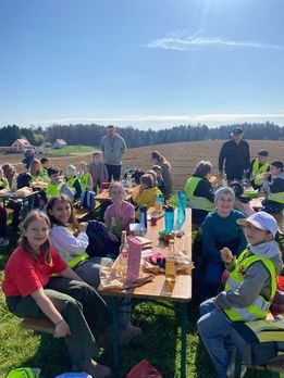 Eine Gruppe von Menschen in gelben Westen sitzt auf Bänken und Tischen auf einem Grasfeld und genießt eine Mahlzeit unter einem klaren blauen Himmel. Einige Erwachsene stehen hinter ihnen. In der Ferne gibt es Bäume und Gebäude.