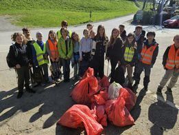 Eine Gruppe von Menschen in Sicherheitswesten steht neben Säcken mit gesammeltem Müll, mit einem grün bewachsenen Hügel und einer Straße im Hintergrund.