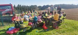Eine Gruppe von Kindern sitzt an Picknicktischen auf einem Feld mit einem roten LKW in der Nähe. Sie tragen Warnwesten und scheinen ein Picknick zu machen.
