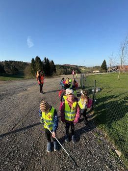 Kinder in Warnwesten laufen auf einem Kiesweg, eines hält einen Stock, umgeben von einem eingezäunten Rasenbereich und entfernten Bäumen und einem Gebäude.