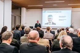 Eine Konferenz findet in einem Raum mit einer Projektionsleinwand statt, auf der ein Mann abgebildet ist. Der Mann steht vor einem Podium mit einem Laptop, und das Publikum sitzt vor ihm.