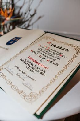 Ein offenes Buch mit grünem und goldenem Rand zeigt ein Zertifikat in deutscher Sprache, das Michael Fuchs-Woringer für seine Beiträge zur russisch-deutschen Freundschaft verliehen wurde. Das Zertifikat ist unterzeichnet und datiert auf 2013.