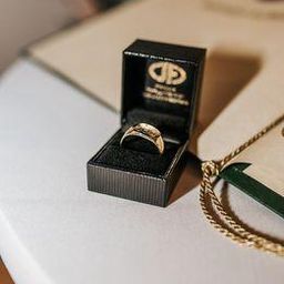 Ein schwarzes Schmuckkästchen auf einem weißen Tisch enthält einen goldenen Ring. Eine goldene Kette liegt neben der Schachtel.