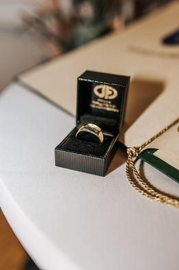 Ein schwarzes Schmuckkästchen auf einem weißen Tisch enthält einen goldenen Ring. Eine goldene Kette liegt neben der Schachtel.