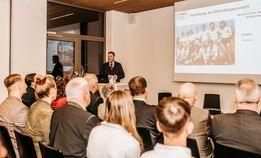 Ein Mann steht vor einer Projektionsleinwand in einem Besprechungsraum. Ein Publikum sitzt in Stühlen vor ihm und hört zu.