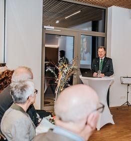 Ein Mann steht an einem Podium vor einem Publikum. Er hält ein Glas und einen Laptop. Eine Glastür mit einer Person auf dem Bildschirm befindet sich hinter ihm.