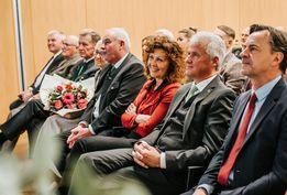 Eine Gruppe älterer Menschen sitzt zusammen auf einer Bank, einige tragen Krawatten und Anzüge. Eine Frau sitzt mit verschränkten Armen.