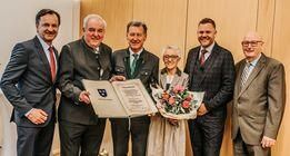 Sechs Männer und eine Frau halten ein Zertifikat und Blumen. Sie stehen eng beieinander, lächeln und scheinen für ein Foto zu posieren.