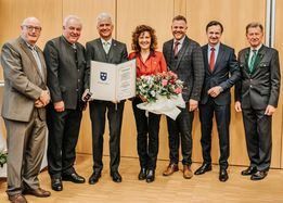 Sieben Personen in formeller Kleidung stehen zusammen und lächeln für ein Foto. Eine Frau in der Mitte hält ein Zertifikat und Blumen. Der Ort scheint eine formelle Veranstaltung zu sein.