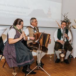 Drei Musiker spielen traditionelle Instrumente; eine Frau spielt Flöte, ein Mann Akkordeon und ein anderer Mann Klarinette, alle sitzen auf Stühlen vor einer Projektionsleinwand.