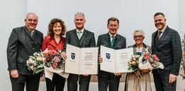 Eine Gruppe formell gekleideter Personen steht zusammen und hält Urkunden und Blumensträuße. Sie scheinen ein bedeutendes Ereignis oder eine Leistung zu feiern.