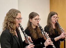 Drei Frauen spielen Klarinette in einem Raum. Sie sind in Schwarz gekleidet, tragen eine Brille und haben lange Haare. Die Frau auf der linken Seite hat lockiges Haar und eine schwarze Jacke. Die Frau in der Mitte hat glattes Haar und trägt eine Halskette. Die Frau auf der rechten Seite hat glattes Haar und trägt einen Armreif.