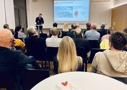 Ein Mann spricht an einem Podium vor einem sitzenden Publikum in einem Konferenzraum mit einer Projektionsleinwand hinter ihm.