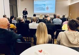 Ein Mann spricht an einem Podium vor einem sitzenden Publikum in einem Konferenzraum mit einer Projektionsleinwand hinter ihm.