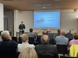 Ein Mann steht an einem Podium und hält eine Präsentation vor einer Leinwand mit einem Projektor. Das Publikum besteht aus Erwachsenen in formeller Kleidung, die auf Stühlen sitzen.