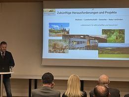 Ein Mann hält eine Präsentation vor einer Projektionsleinwand mit Bildern und Text über zukünftige Herausforderungen und Projekte. Das Publikum sitzt und hört zu.