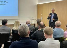 Ein Mann hält eine Präsentation für ein sitzendes Publikum in einem Konferenzraum. Der Mann steht vor einer Projektionsleinwand, und das Publikum hört aufmerksam zu.