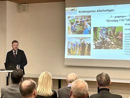 Ein Mann in Anzug präsentiert vor einer Projektionsleinwand mit 'Kindergarten Alleheiligen' und Bildern von Kindern. Mehrere Leute in Anzügen sitzen vor ihm und hören zu.