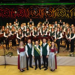 Ein großes Orchester mit verschiedenen Instrumenten posiert für ein Foto auf der Bühne, mit Blumendekorationen im Vordergrund und einem gemusterten Hintergrund.