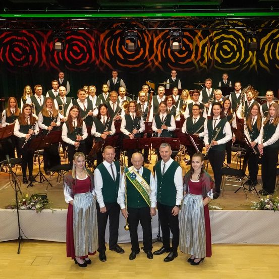 Ein großes Orchester mit verschiedenen Instrumenten posiert für ein Foto auf der Bühne, mit Blumendekorationen im Vordergrund und einem gemusterten Hintergrund.