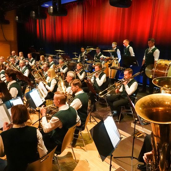 Ein Brass-Band spielt auf einer Bühne mit roten Vorhängen. Mehrere Musiker spielen verschiedene Instrumente. Einige sitzen, andere stehen. Es gibt Blechblasinstrumente, Trommeln und einen Notenständer.