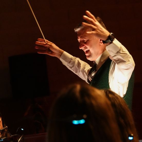 Ein Mann, möglicherweise ein Dirigent, leitet eine musikalische Aufführung mit einem Taktstock. Er trägt eine Uhr, eine grüne Weste und eine Krawatte. Eine Frau beobachtet ihn.