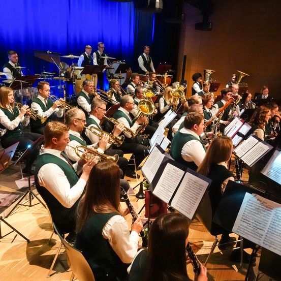 Ein großes Orchester spielt auf der Bühne, mit Musikern, die Trompeten, Posaunen und andere Instrumente spielen. Die Musiker sitzen vor Notenständern, mit einem blauen Vorhang als Hintergrund.