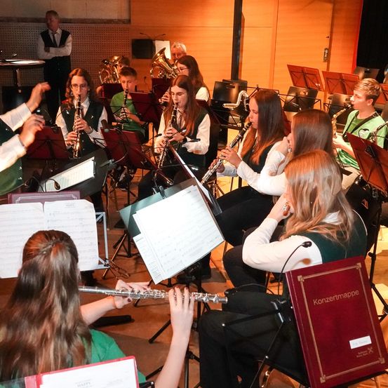 Eine Gruppe junger Musiker sitzt und spielt verschiedene Instrumente in einem Raum. Einige spielen Flöte, Klarinette und Trompete. Sie haben Notenblätter vor sich und einen Notenständer in der Nähe.