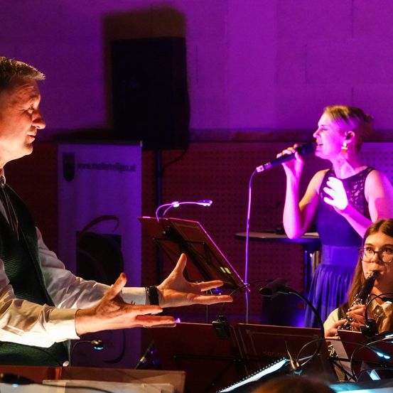 Ein Musiker in einer grünen Weste dirigiert ein Orchester, eine Frau singt in ein Mikrofon und eine andere spielt Klarinette. Die Szene ist in lila Licht getaucht.