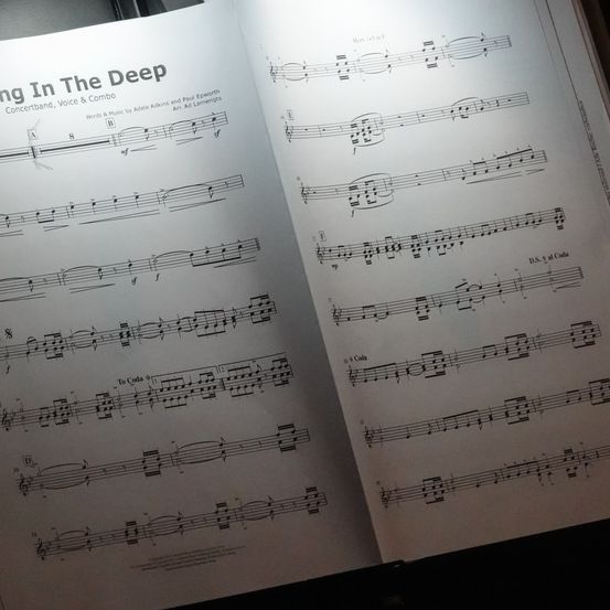 Ein offenes Musikbuch zeigt Noten für ein Stück mit dem Titel 'Sing In The Deep' von Adele Adkins und Paul Epworth. Die Noten sind für Konzertband, Stimme und Combo arrangiert.