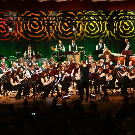 Ein Orchester spielt auf der Bühne vor einem großen Publikum, mit Trompeten, Trommeln und Flöten. Der Hintergrund hat leuchtende Wirbel, und die Künstler sind in passenden grünen Westen gekleidet.