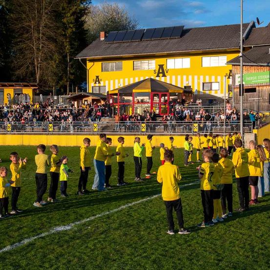 Eine Gruppe von Kindern in gelben Trikots steht auf einem Fußballfeld, mit einer Menge in den Tribünen. Dahinter befindet sich ein gelbes Gebäude mit Solarmodulen auf dem Dach.