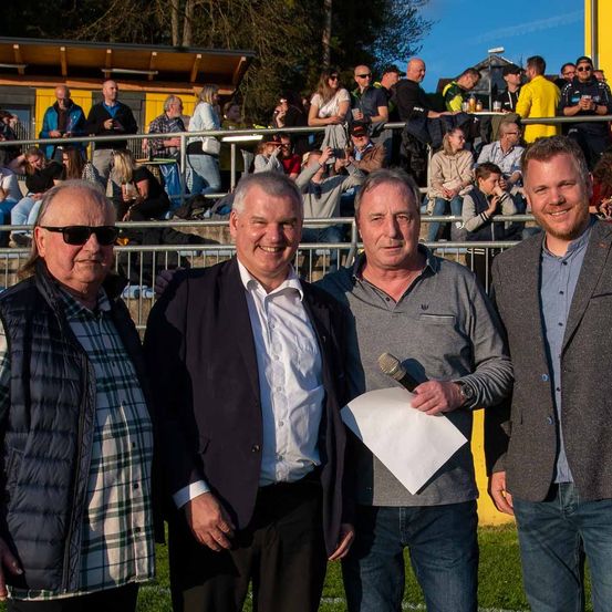 Vier Männer stehen auf einem Sportfeld. Sie lächeln und scheinen für ein Foto zu posieren. Der Mann in der Mitte hält ein Mikrofon und ein Papier. Hinter ihnen sitzen viele Menschen auf den Tribünen. Einige halten Tassen und machen Fotos.