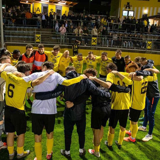 Eine Fußballmannschaft versammelt sich auf dem Spielfeld, während eine Menge auf den Tribünen zuschaut. Alle tragen gelbe Trikots außer einem Spieler in Weiß.