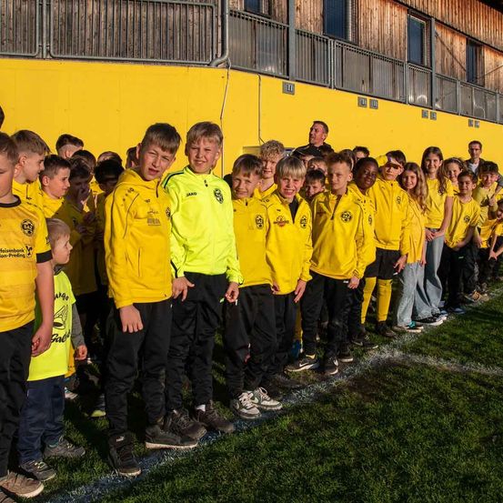 Eine Gruppe von Kindern in gelben Trikots steht in einer Reihe, wobei ein Junge in einem gelben Jackett hervorsticht. Sie befinden sich auf einem Fußballplatz, mit einer gelben Wand und einem Gebäude im Hintergrund.