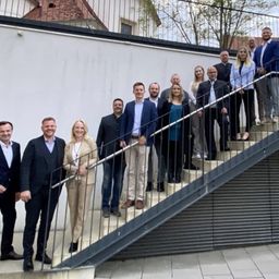 Eine Gruppe formell gekleideter Personen steht auf einer Treppe mit Metallgeländern und lächelt für ein Foto vor einer weißen Wand mit Bäumen im Hintergrund.