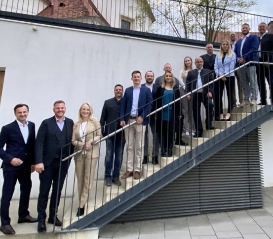 Eine Gruppe formell gekleideter Personen steht auf einer Treppe mit Metallgeländern und lächelt für ein Foto vor einer weißen Wand mit Bäumen im Hintergrund.