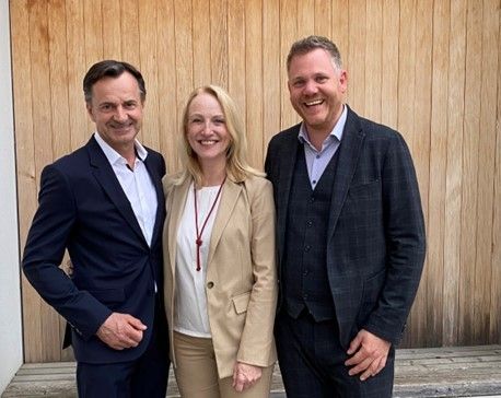 Drei Menschen stehen lächelnd vor einer Holzwand. Der Mann links trägt einen dunklen Anzug. Die Frau in der Mitte trägt einen beigen Blazer. Der Mann rechts trägt einen dunklen Mantel.